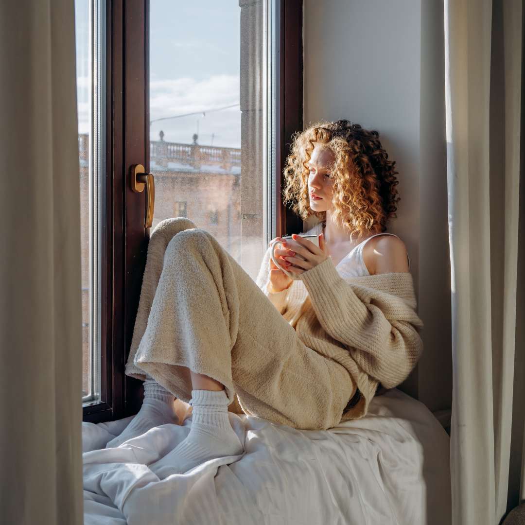 Kobieta siedząca na parapecie z kubkiem parującej herbaty, patrząca przez okno. Estetyczna ilustracja równowagi między biznesem a życiem prywatnym (work-life balance) w modelu FlowSync.